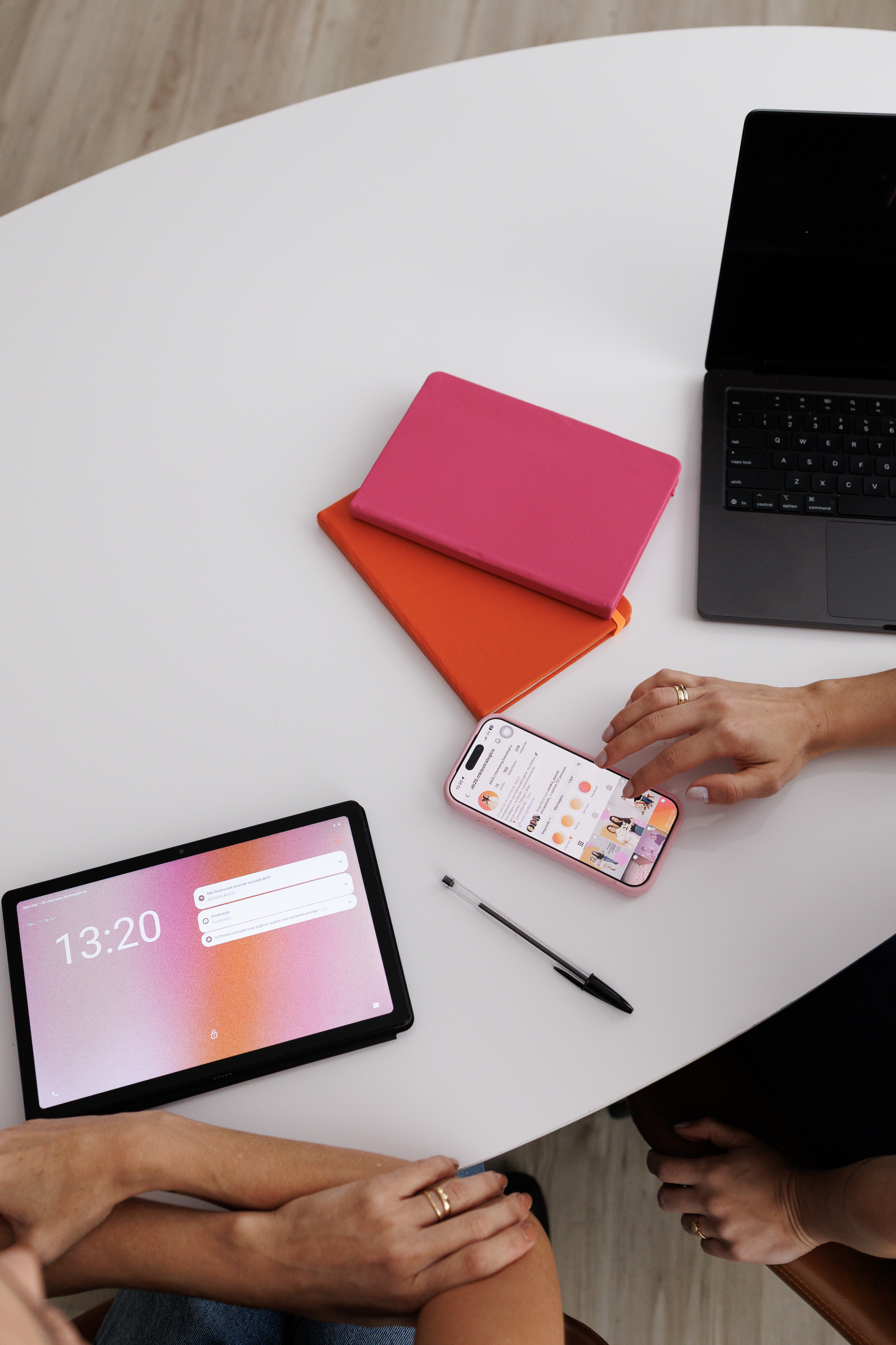 Mesa com cadernos rosa e laranja e celular mostrando o perfil da AB26 no Instagram